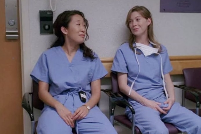 Two women in blue scrubs sitting on a bench in a hospital, looking at each other.