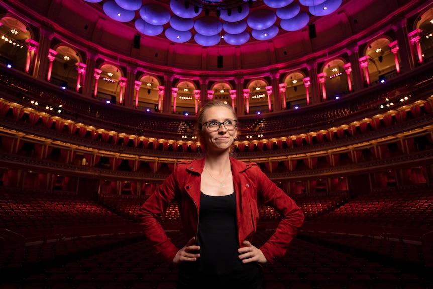 Anna Lapwood on stage at the Royal Albert Hall with seating behind her. Her hands are on hips and she smiles looking off camera.