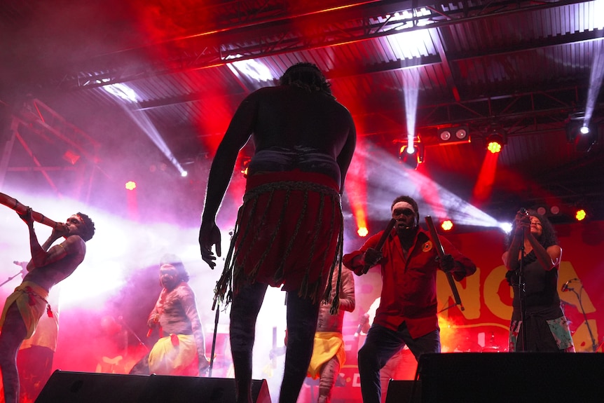 A photo taken from behind shows a man in a lion cloth, in shaows, red strobe lighting, Indigenous men play the didgeridoo.