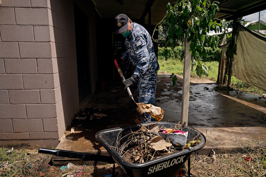 A man in a camoflauge uniform shovels rubbish into a wheelbarrow.