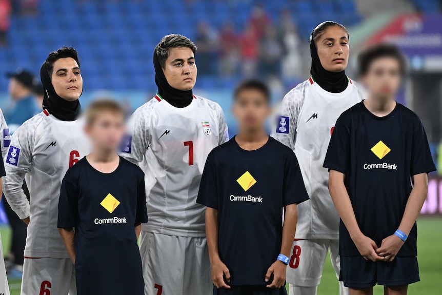 Three members of the Iran women's football team line up listening to their national anthem before a game.