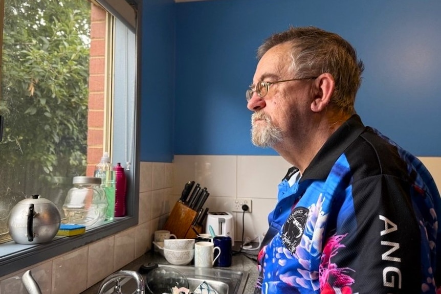 A middle aged white man with short grey hair and glasses looking out a kitchen window