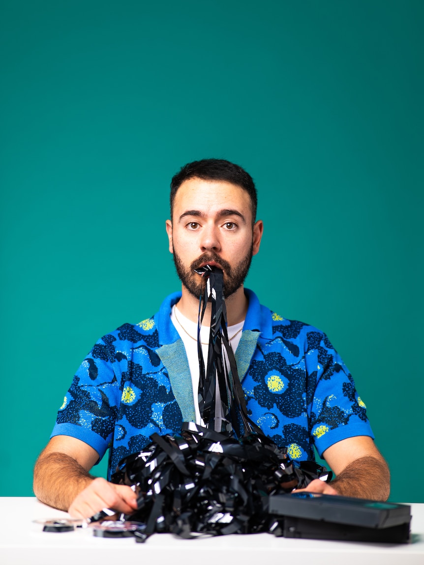 Anesti Danelis, Greek Canadian, early 30s, sits at a table, mouth full of the unwound tape from video tapes.