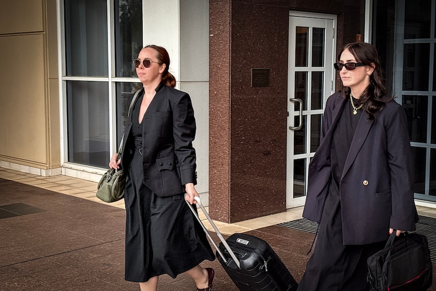 Two white women in black dress suits walking out of court building, sunglasses on, one hold suitcase.