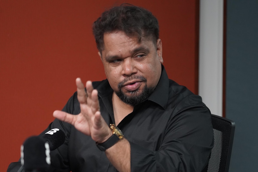 A man mid-gesture talking into a microphone and looking over his shoulder.