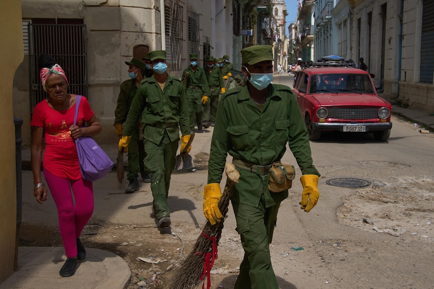Cuban soldiers in green clothing and yellow gloves wearing blue face masks walking on a road next to an old red car.