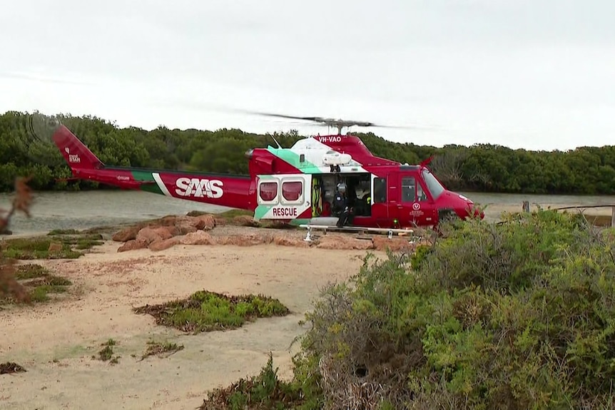 A red and white helicopter with an open side door lands on a sandy area