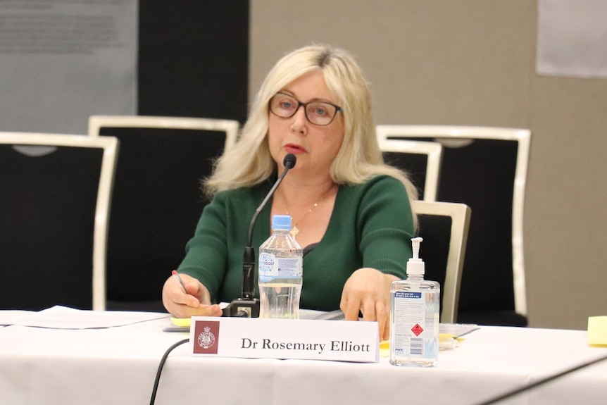 A woman with long blonde hair and a dark green top sits at a desk behind a microphone.