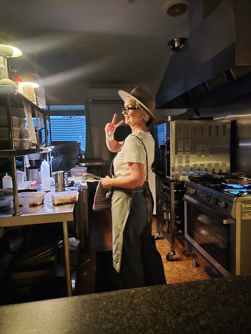 Woman throwing up peace sign while standing in commercial kitchen, big smile