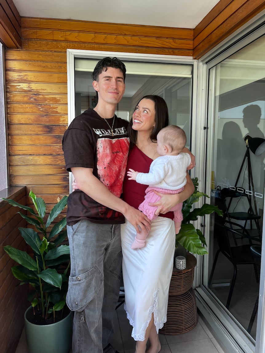 Madeleine Edwards holds her baby Carmien on her hip and poses with her partner Matt on a balcony.