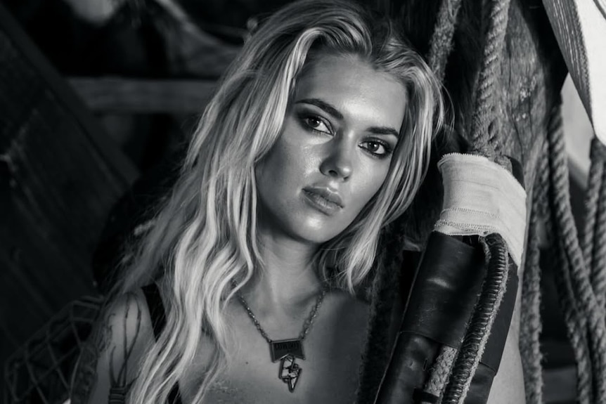 A woman with long blonde hair and a black bra holds onto a rope.