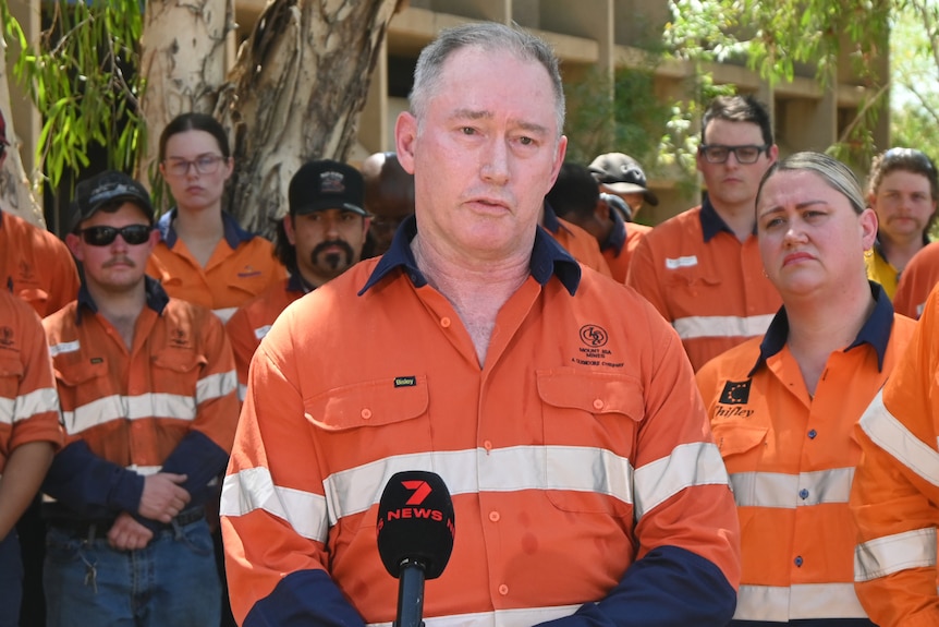 A man in glencore mining branded orange hi-viz overalls addresses a press conference. 
