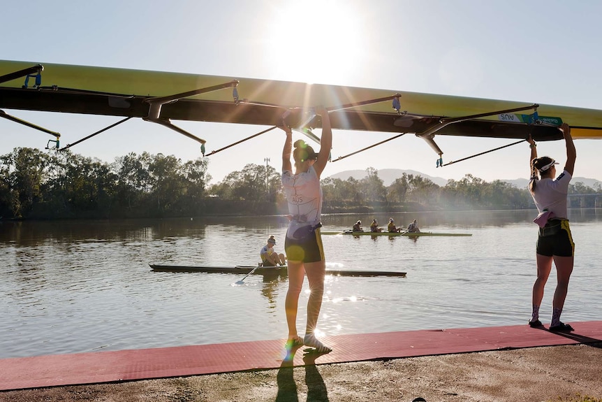 Two women hold a rowing boat above their heads as they prepare to place it in the water. Other rowers in background on river.