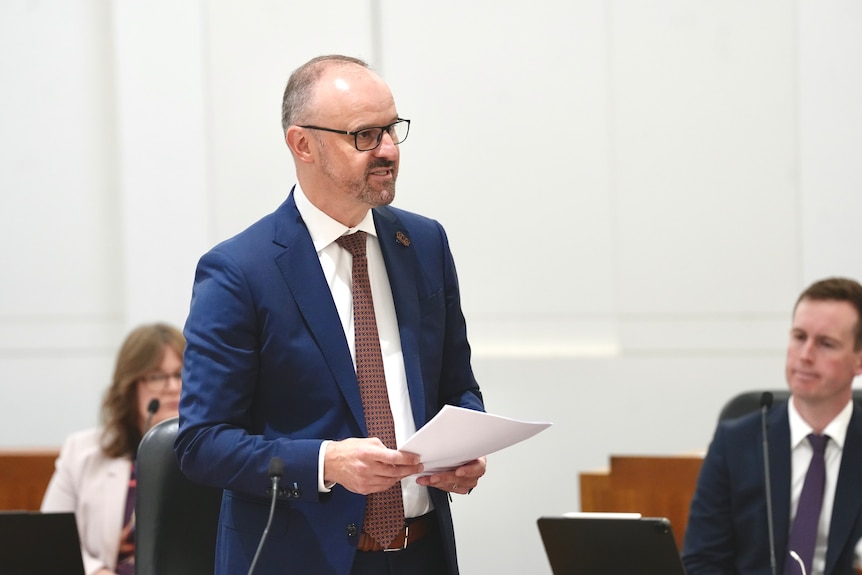 A man in a navy blue suits stands in a room with red seats speaking seriously.