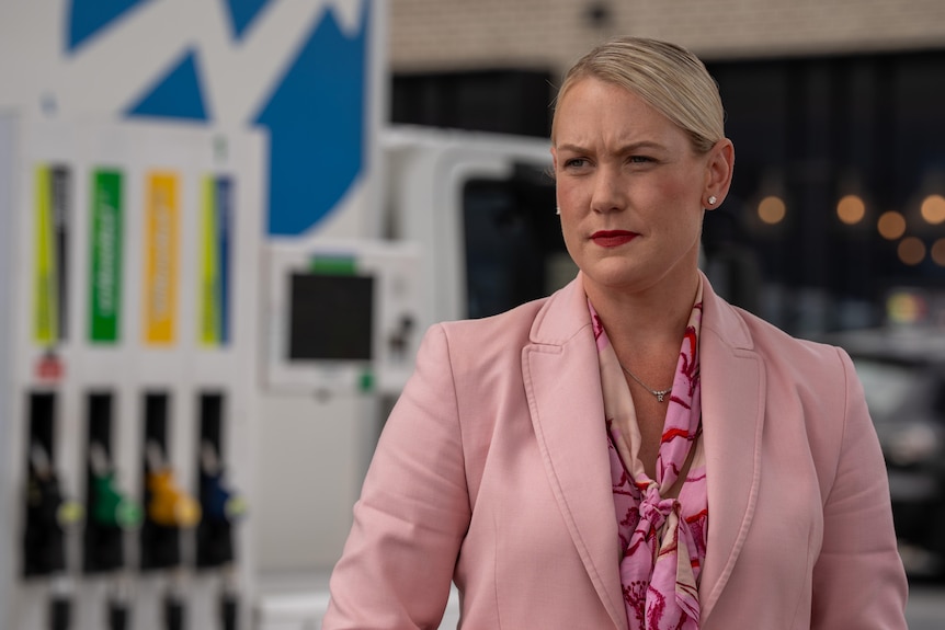 A woman wearing a pink suit and a serious expression stands in front of petrol pumps