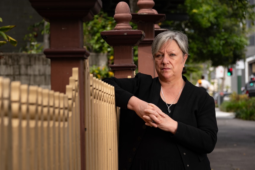 A middle-aged woman with short hair looks solemn as she leans against a fence on a city street.