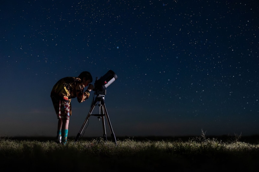 A silhouetted person looks into a telescope on a dark night.