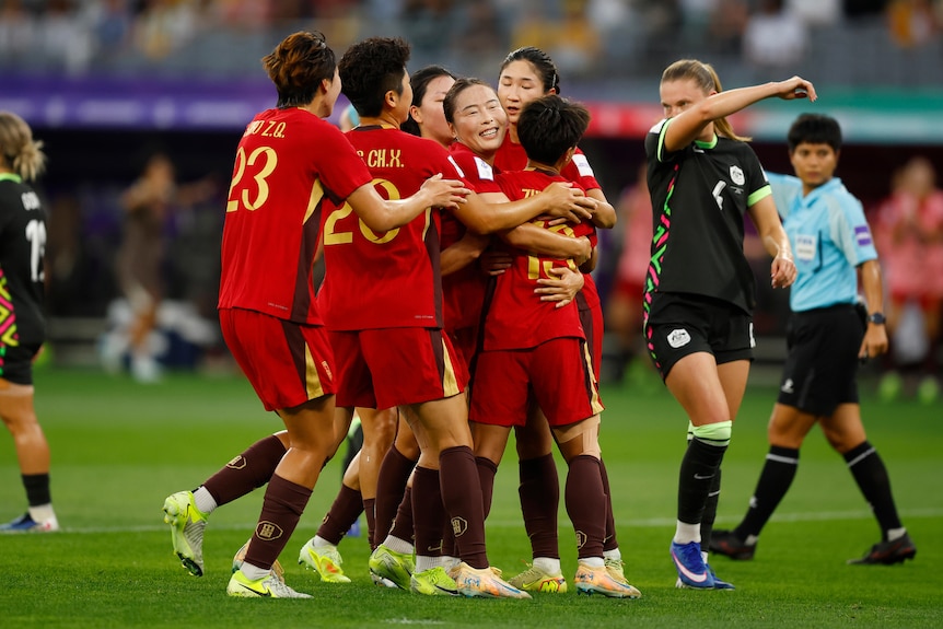 A group of soccer players in red celebrate a goal with player in green behind them