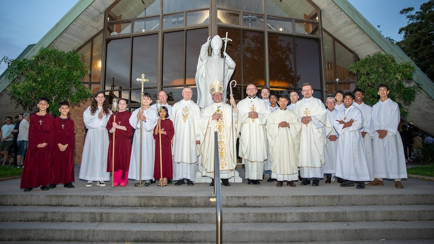 more than a dozen church stuff in white robes, with a church behind them