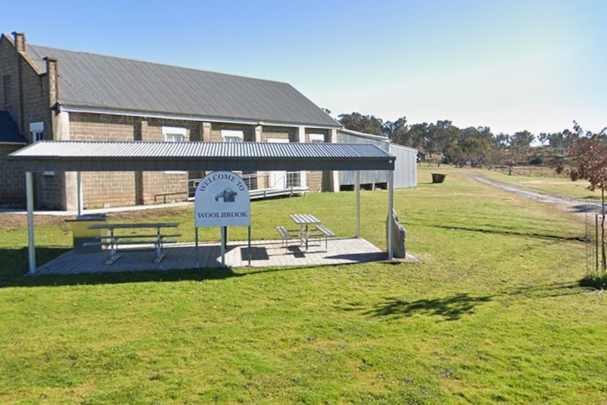 grassed area with undercover picnic tables. sign says 'welcome to woolbrook', behind seating area is brick community hall.