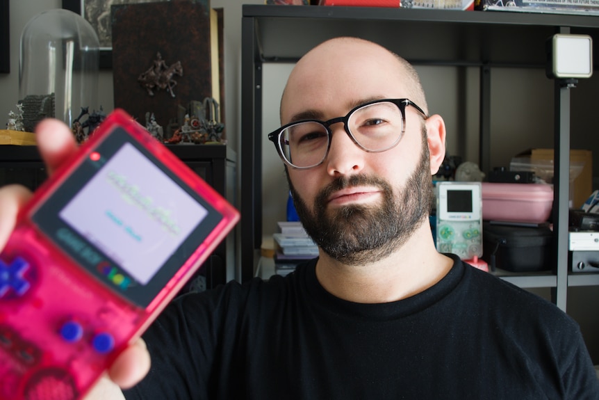 A man with glasses and a beard holds a gameboy towards the camera which is pink with a backlit screen
