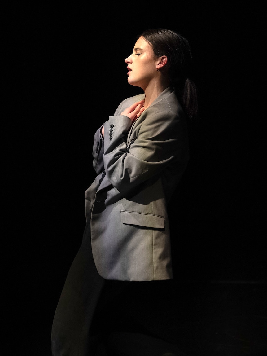 A dark-haired woman in a coat strikes a pose while performing on a darkened stage.