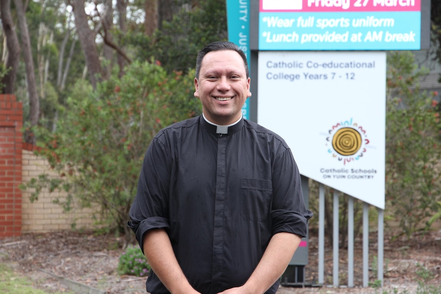 A Catholic minister standing in front of a sign.