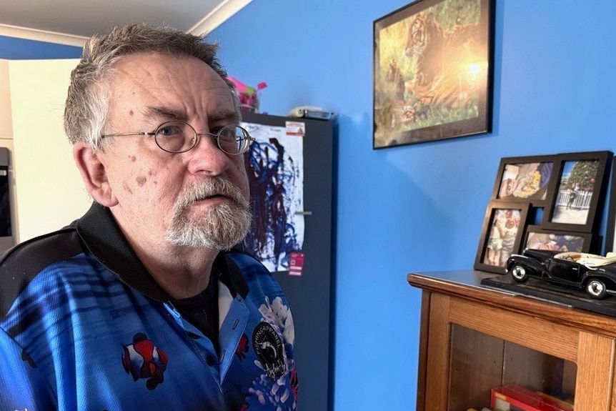A middle aged white man with short grey hair and glasses standing by a blue wall