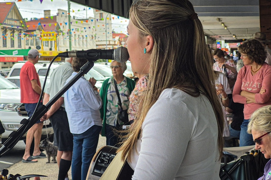 A woman sings into a guitar as a street full of people bustles in front of her. 