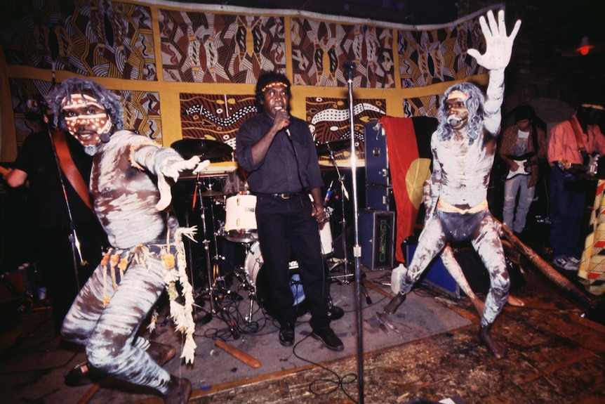 Two men in Aboriginal traditional body paint mid-dancing on stage while a third man sings into a microphone.