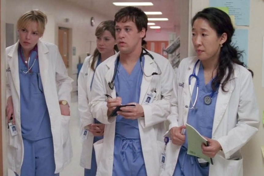 Four people in blue scrubs and white coats stand looking into a room.