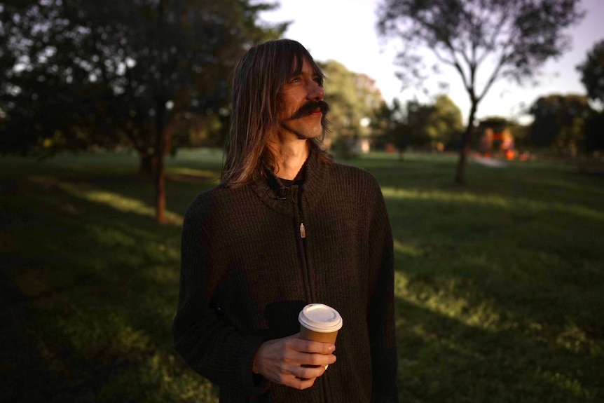 A man with a moustache holds a takeaway coffee cup and looks up.