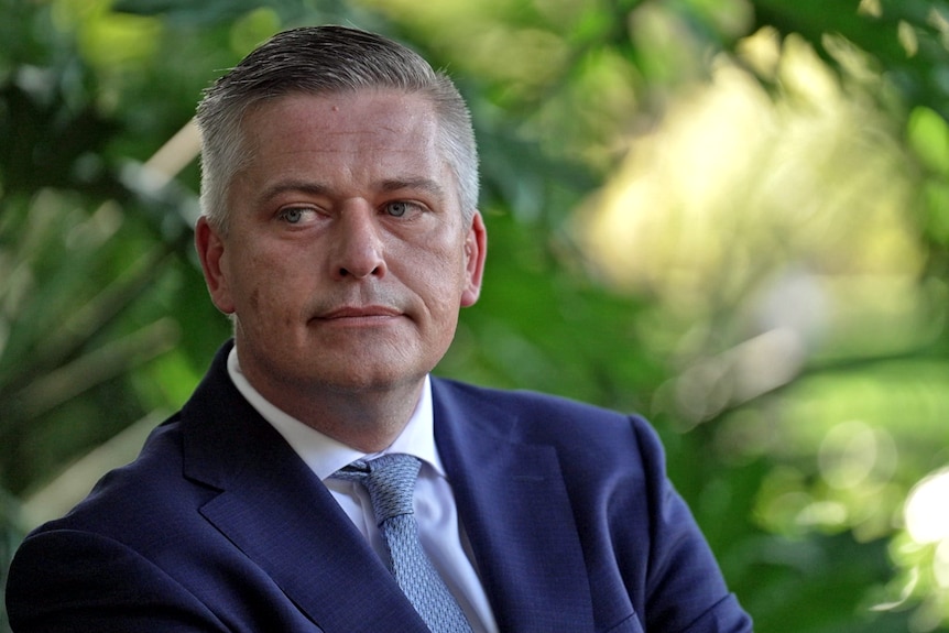 A man in a suit and tie looks to one side, with a neutral expression. Behind him is foliage.