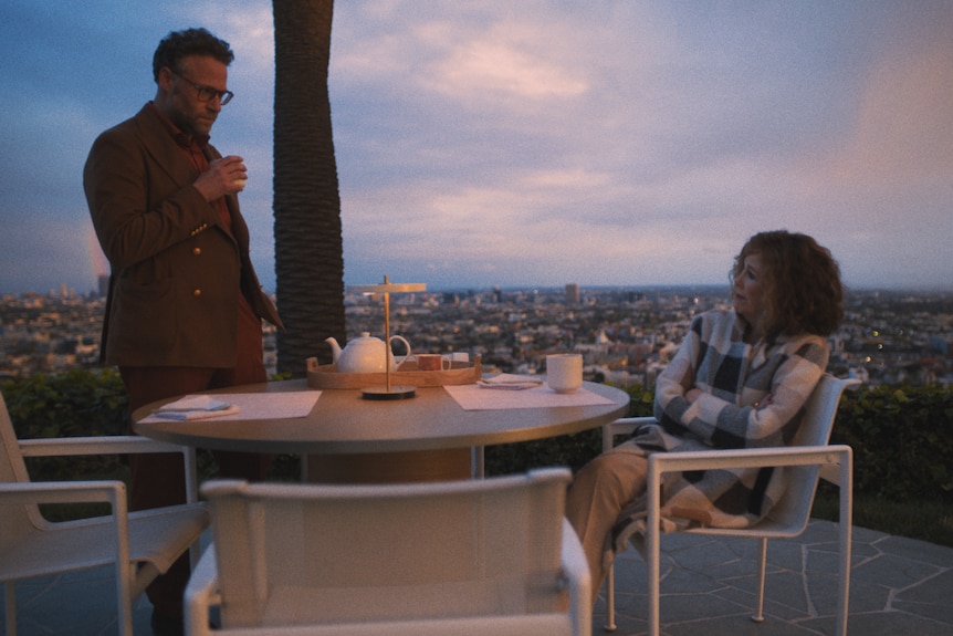 Seth, left, stands around a table with Catherine, right, seated with the LA skyline behind them at dusk mid conversation