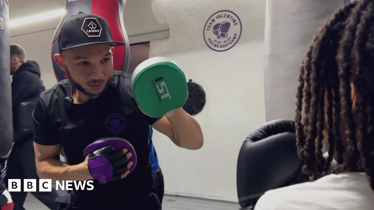 Curtis is wearing a snapback, he has boxing gloves on, and is in his gym. He is wearing a black t-shirt and looks focused.