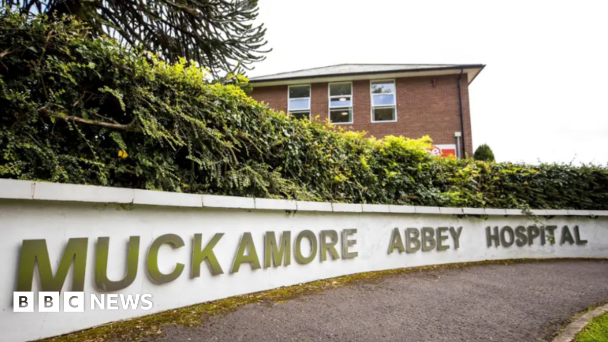 A sign outside a red building that reads: Muckamore Abbey Hospital in gold letters on a white wall.  A hedge is on top of the wall.