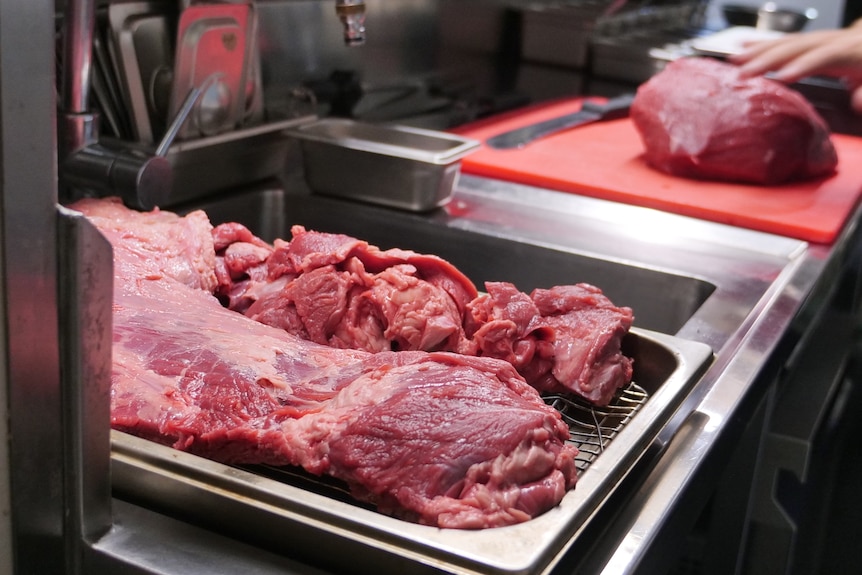 Close up of meet on a tray, with another slab of meat in the background being prepared to be cut.