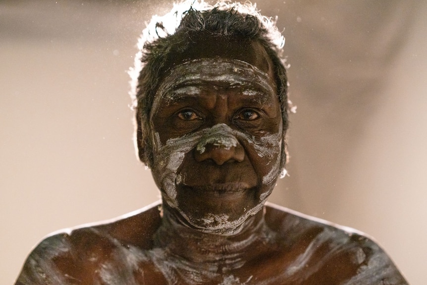 An elderly Aboriginal man covered in traditional white body paint, top half of chest showing, stares at the camera.