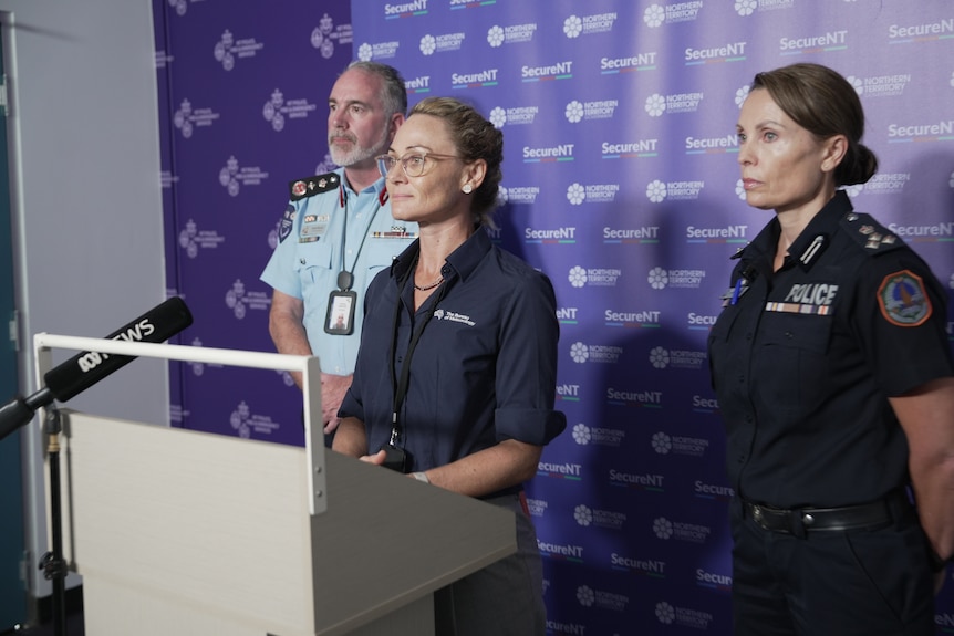 A woman standing at a lectern and in front of a media microphone, flanked by a man and woman.