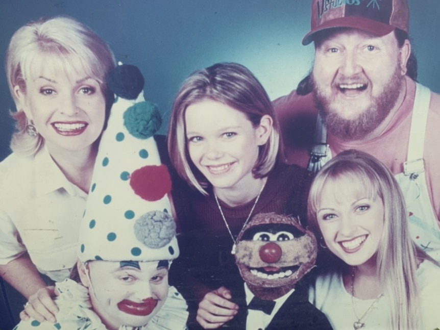 Five people from a TV show in the 1990s smiling at the camera, one is dressed as a clown, and a puppet.