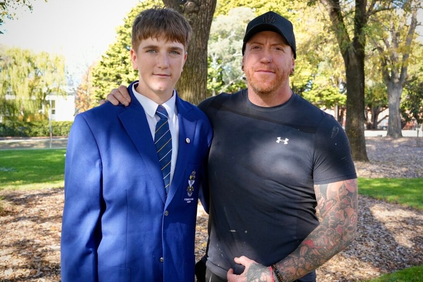 A teenaged boy in a blue blazer stands with a man in a black t-shirt and cap.