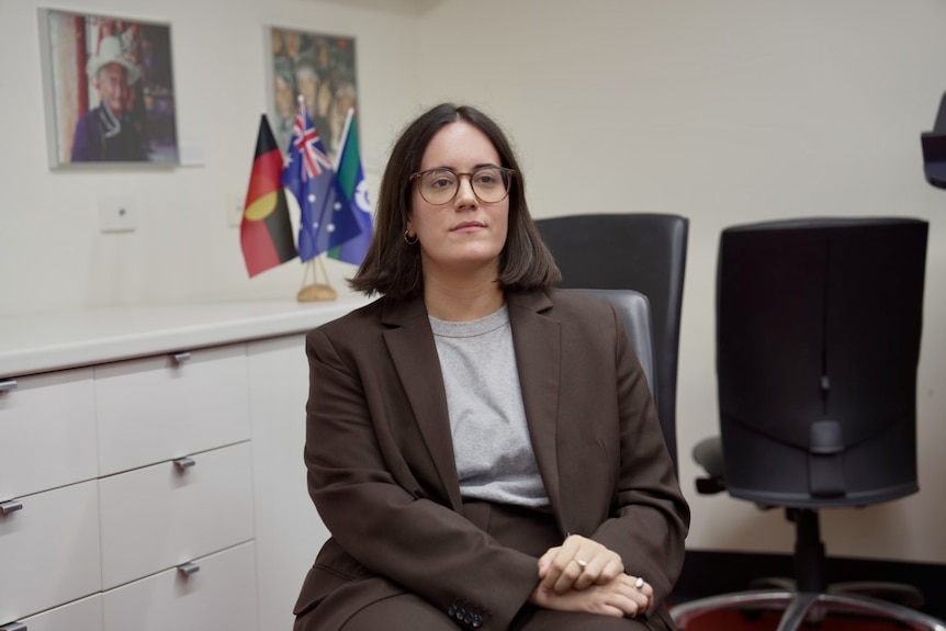 woman with short brown hair and glasses wearing a brown suit sitting in an office with black chairs in the background