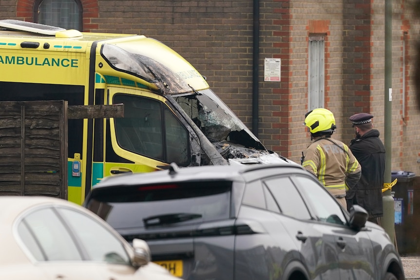 A firefighter and a police officer look at a yellow ambulance which has been damaged by fire. 