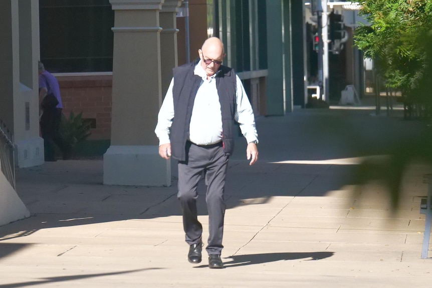 An elderly man in suit pants, a long sleeved striped shirt and jacket walks down a street