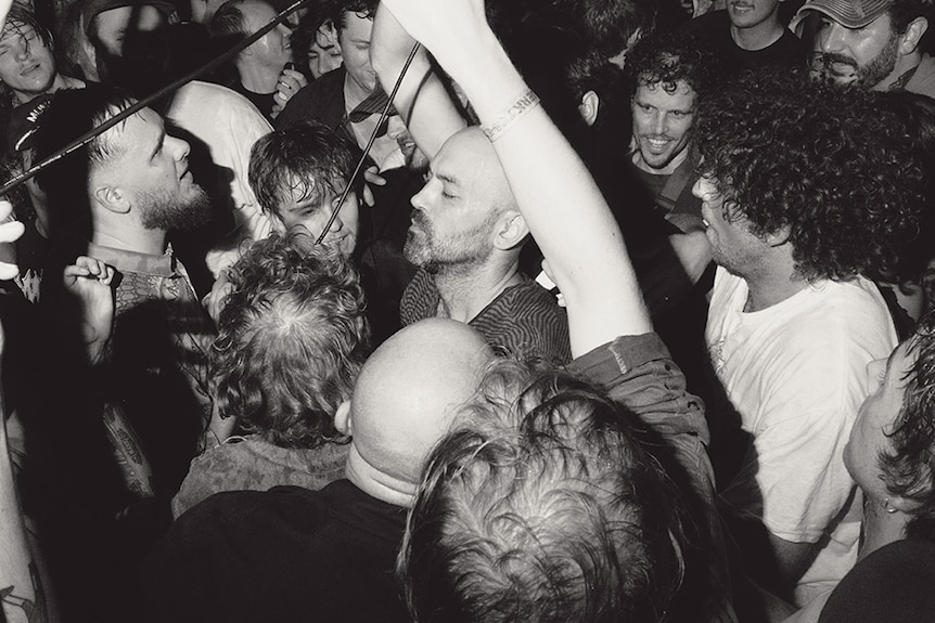 Black and white photo of man with microphone standing in amongst lively audience in an indoor music venue