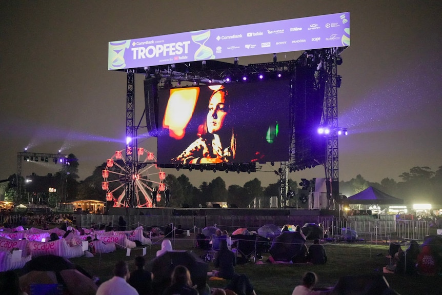 An audience sits outdoors watching a large screen play a short film. 