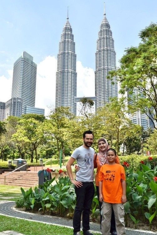 three people stand in a park with the Petronas Tower and a cityscape behind