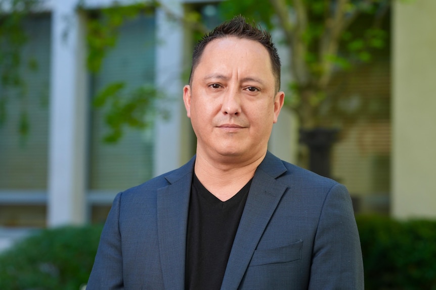 A dark-haired man in a blazer stands near a building lined with a hedge.