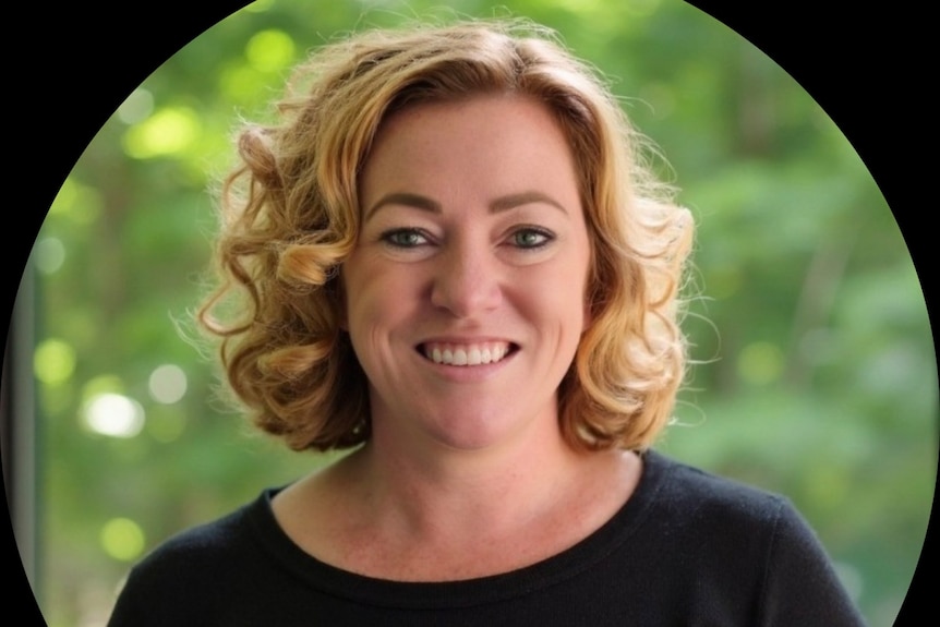 A photo of a woman with blonde curly hair, wearing a black top, with green bushes in the background