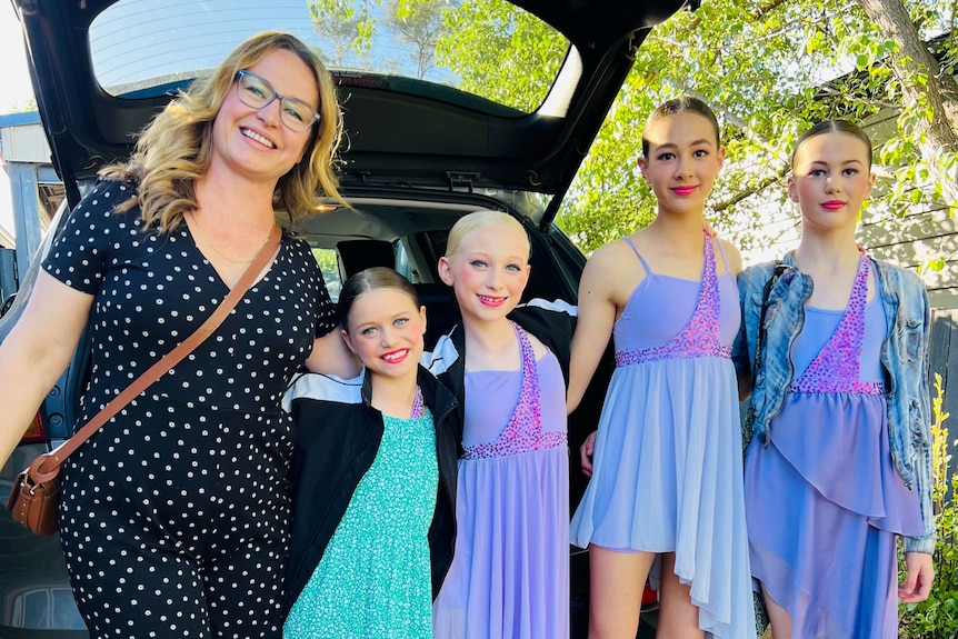 A smiling woman stands beside four girls in face make-up and dance costumes at the back of a car.